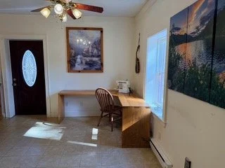 Interior of a room with a wooden desk and chair next to two windows, a painting of a mountain scene on the wall, and a ceiling fan with light fixtures.