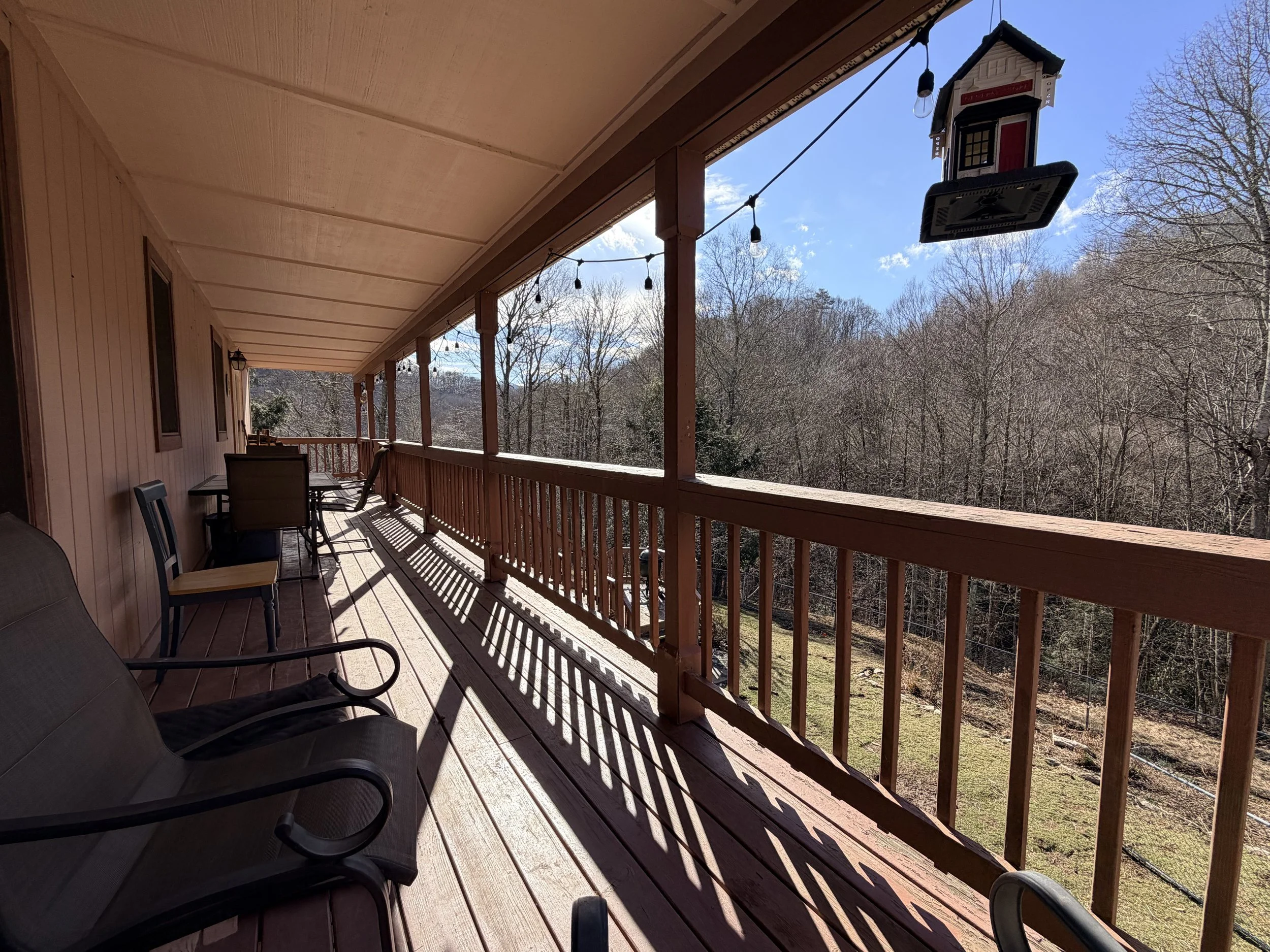 Sunlit wooden deck with chairs, a table, and string lights, overlooking a treeline with leafless trees and a blue sky.