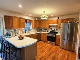 Kitchen with wooden cabinets, stainless steel refrigerator, stove, microwave, and a small island with a bowl of fruit, hardwood flooring, and a ceiling light fixture.