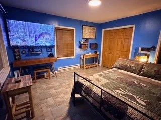 A bedroom with blue walls, a large bed with a patchwork quilt, a wooden dresser, a door, and a wall-mounted flat-screen TV displaying a winter scene with snow. Windows with wooden blinds, and a small table.