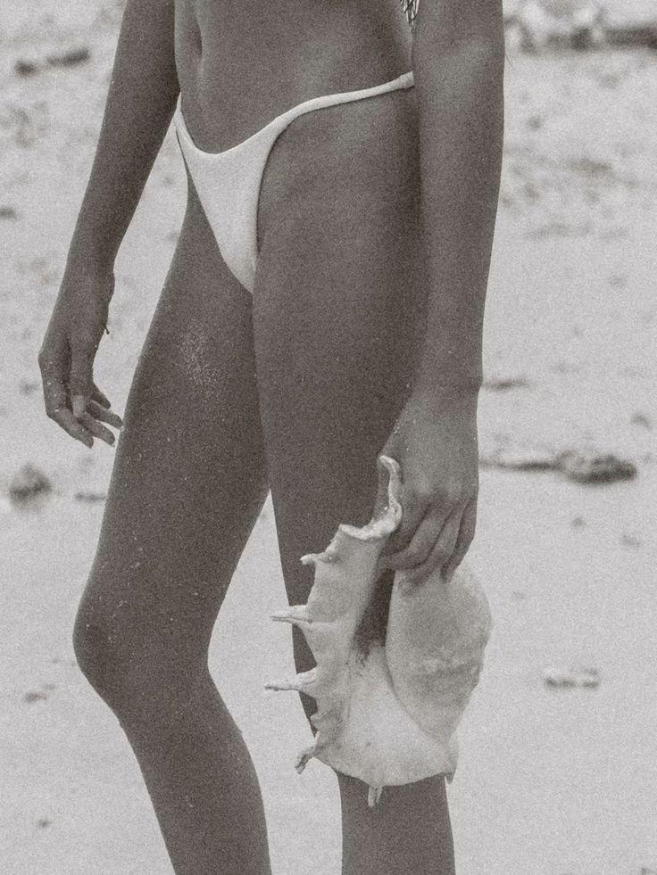 A woman in a bikini holding a large seashell on a beach.