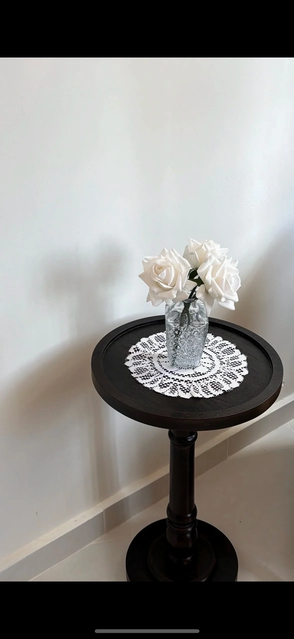 A round black wooden table with a decorative glass vase holding white roses, placed on a white lace doily against a plain white wall.