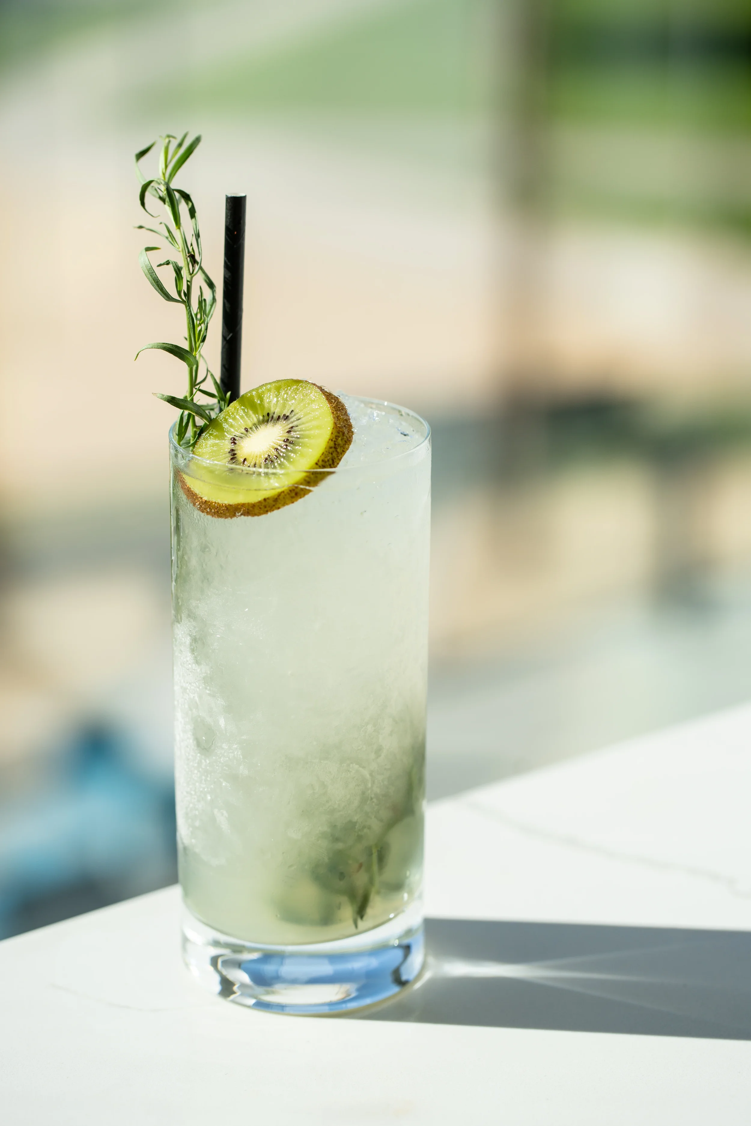 A tall cocktail garnished with kiwi and rosemary on a sunlit surface.