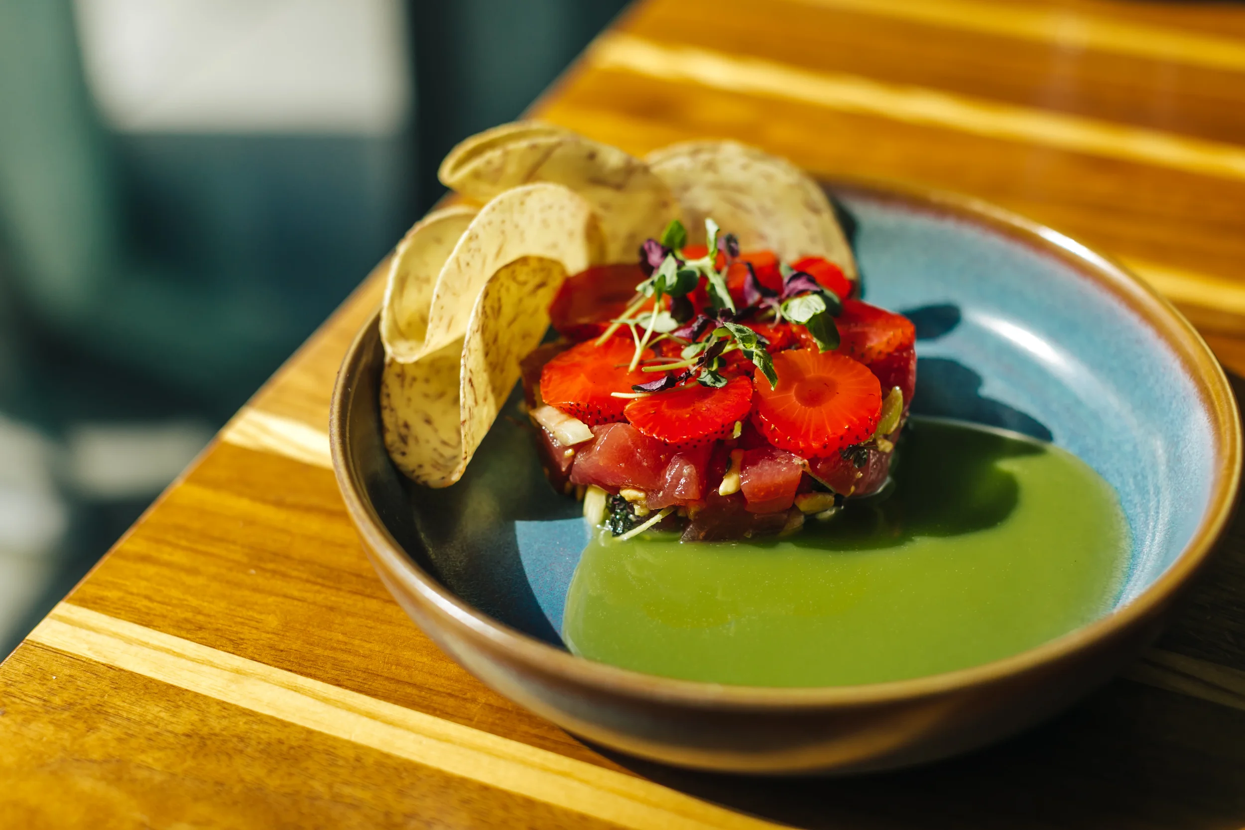 Tuna tartare with strawberries, microgreens, and taro chips on a blue plate with green sauce.