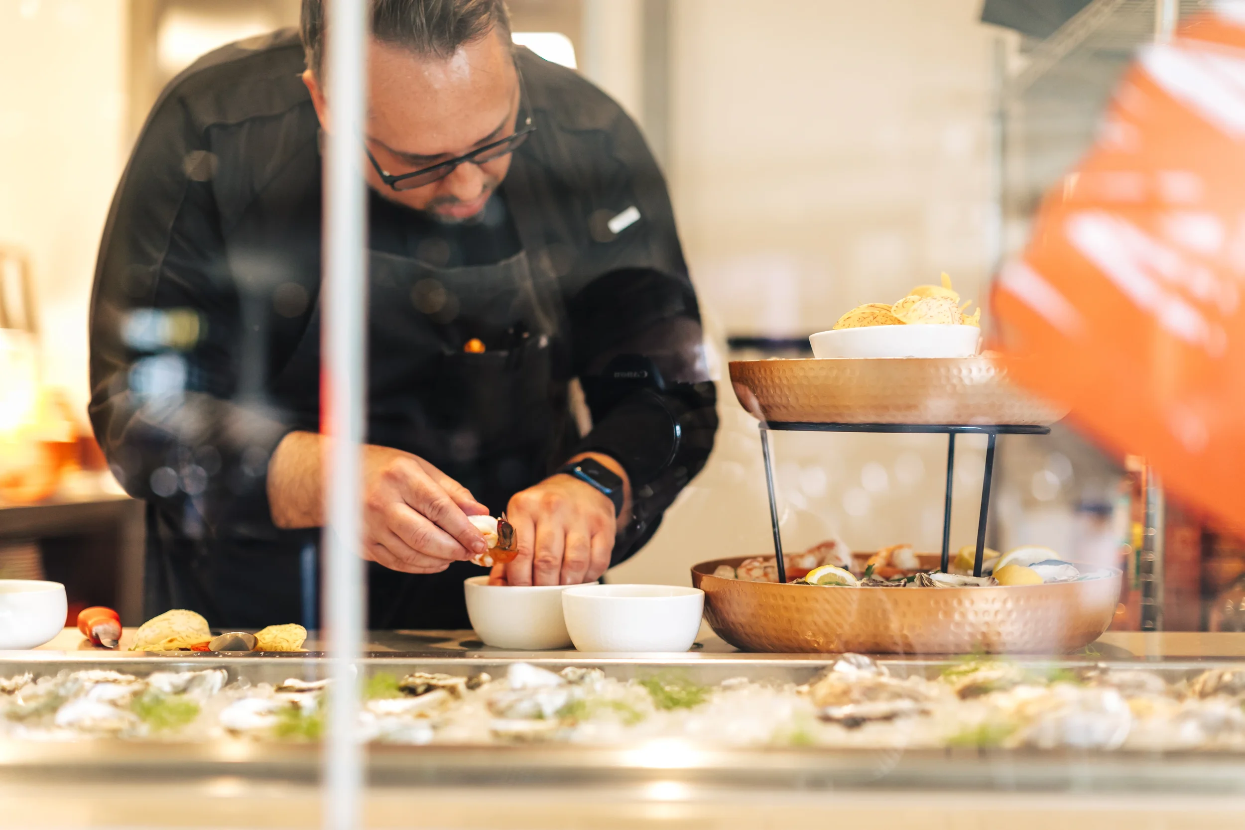 A chef, Franck Tasic, in black prepares seafood at a counter surrounded by ingredients.