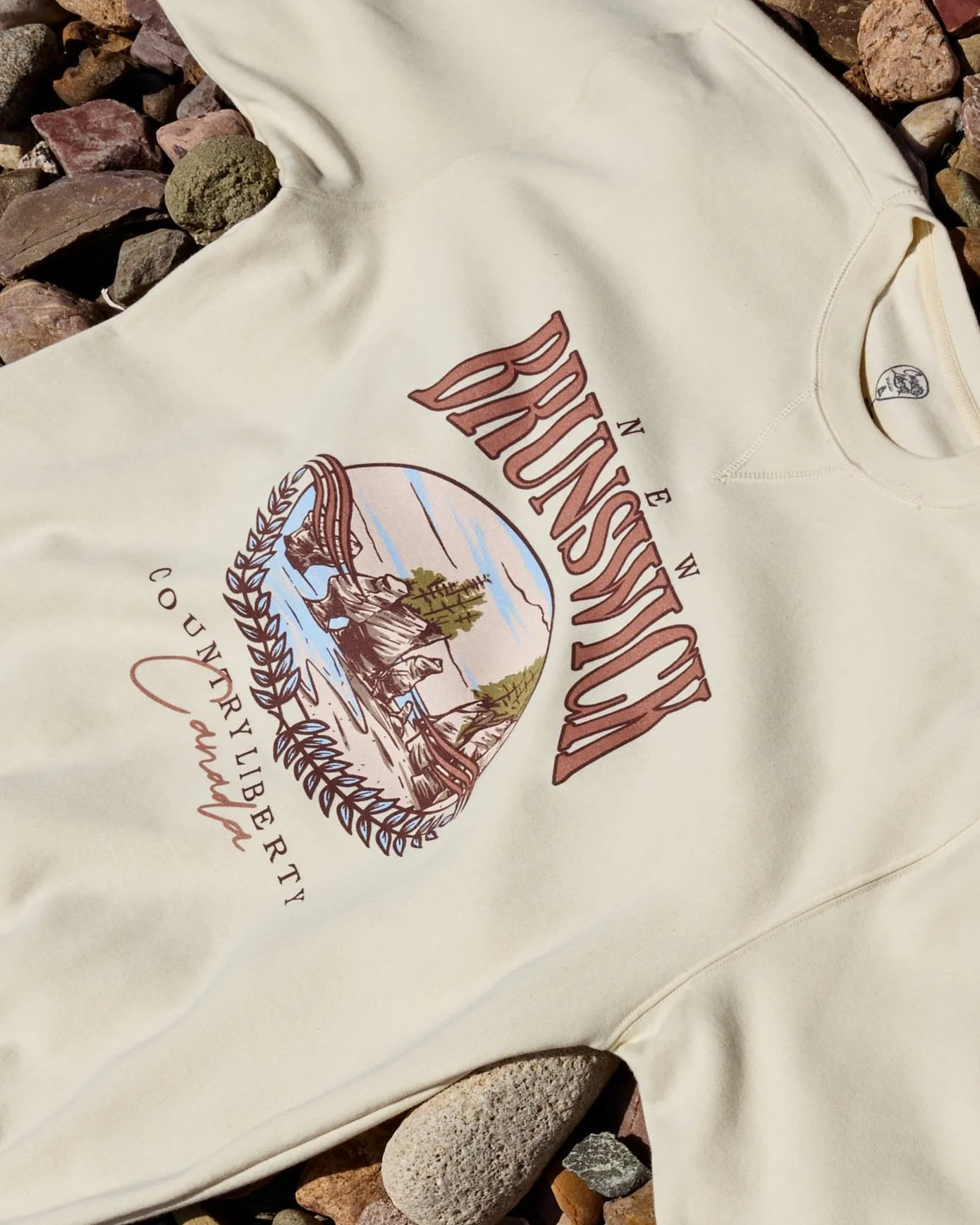 Cream-colored sweatshirt with a mountain, trees, river, and eagle illustration, and the words 'BRUNSWICK COUNTY LIBERTY' on it, placed on rocks.