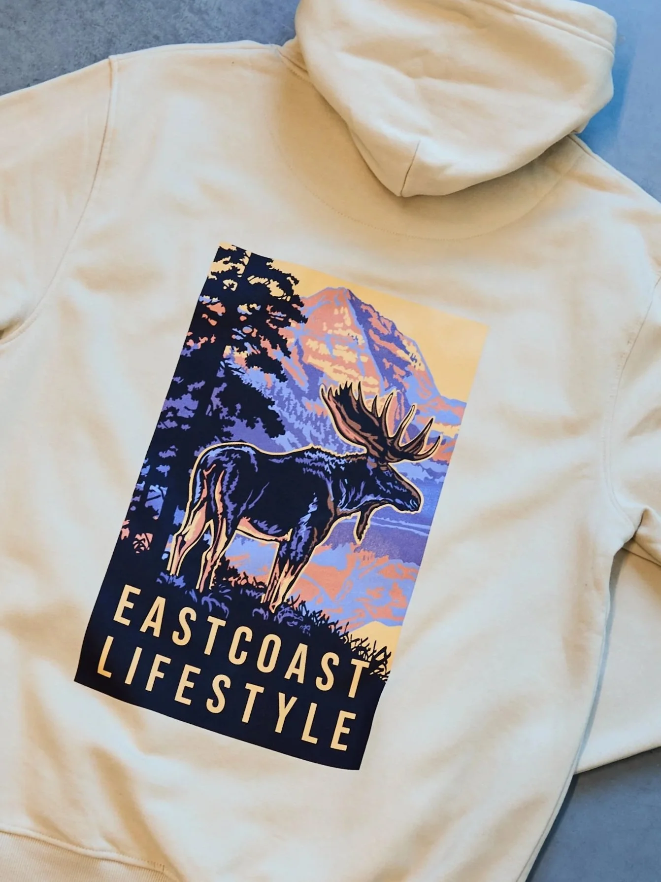 Cream-colored hoodie with a graphic of a moose, trees, and a mountain in pastel colors, with the words "EAST COAST LIFESTYLE" written below.