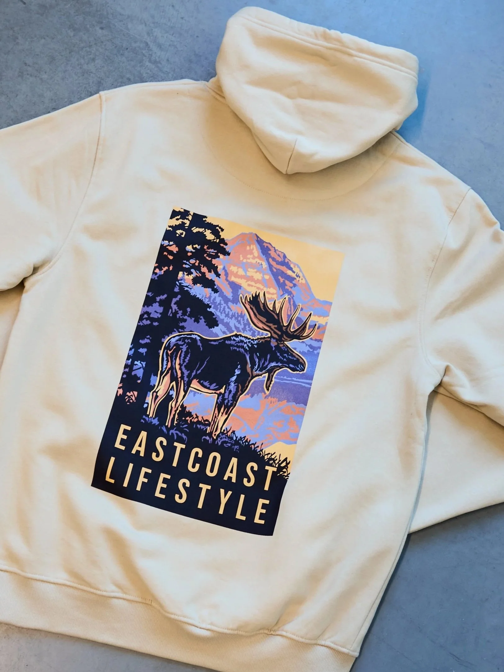 A beige hoodie with a graphic design of a moose in front of a mountain landscape, with the text "EASTCOAST LIFESTYLE" printed below the moose.