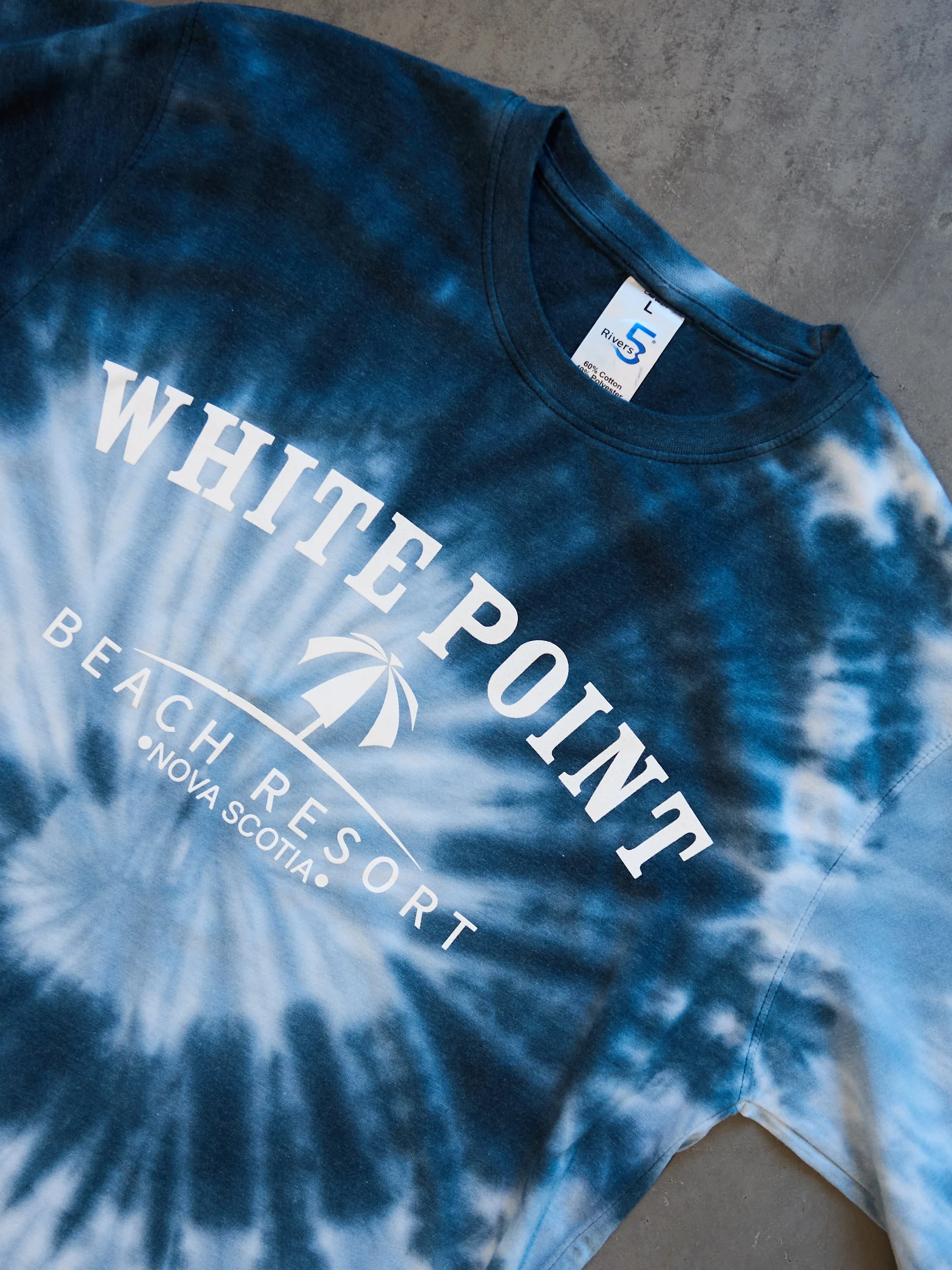 Blue tie-dye t-shirt with white text that reads 'White Point Beach Resort Nova Scotia' and a graphic of a beach umbrella with rays of light.
