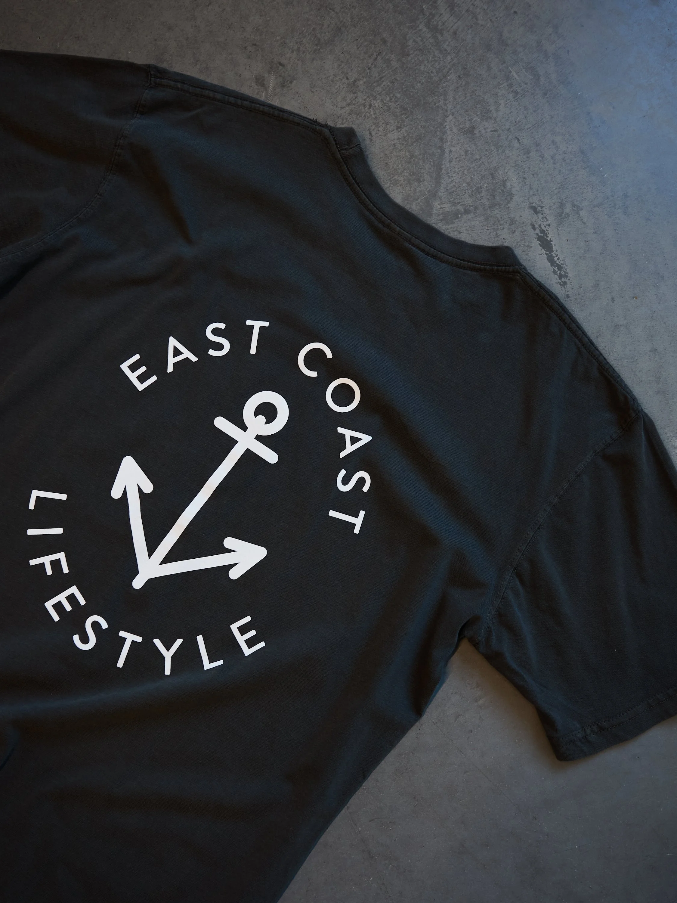 Black T-shirt with white graphic of an anchor and the words 'EAST COAST LIFE STYLE' on a concrete floor.