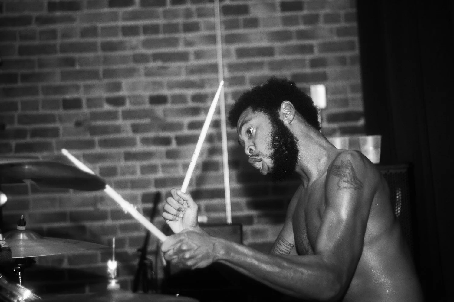 A shirtless man with a tattoo on his right shoulder plays drums in a room with brick walls.