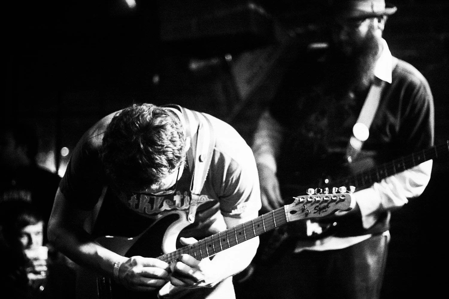 Two guitarists play on stage in a black and white photo, one with head down and the other in profile holding a guitar.