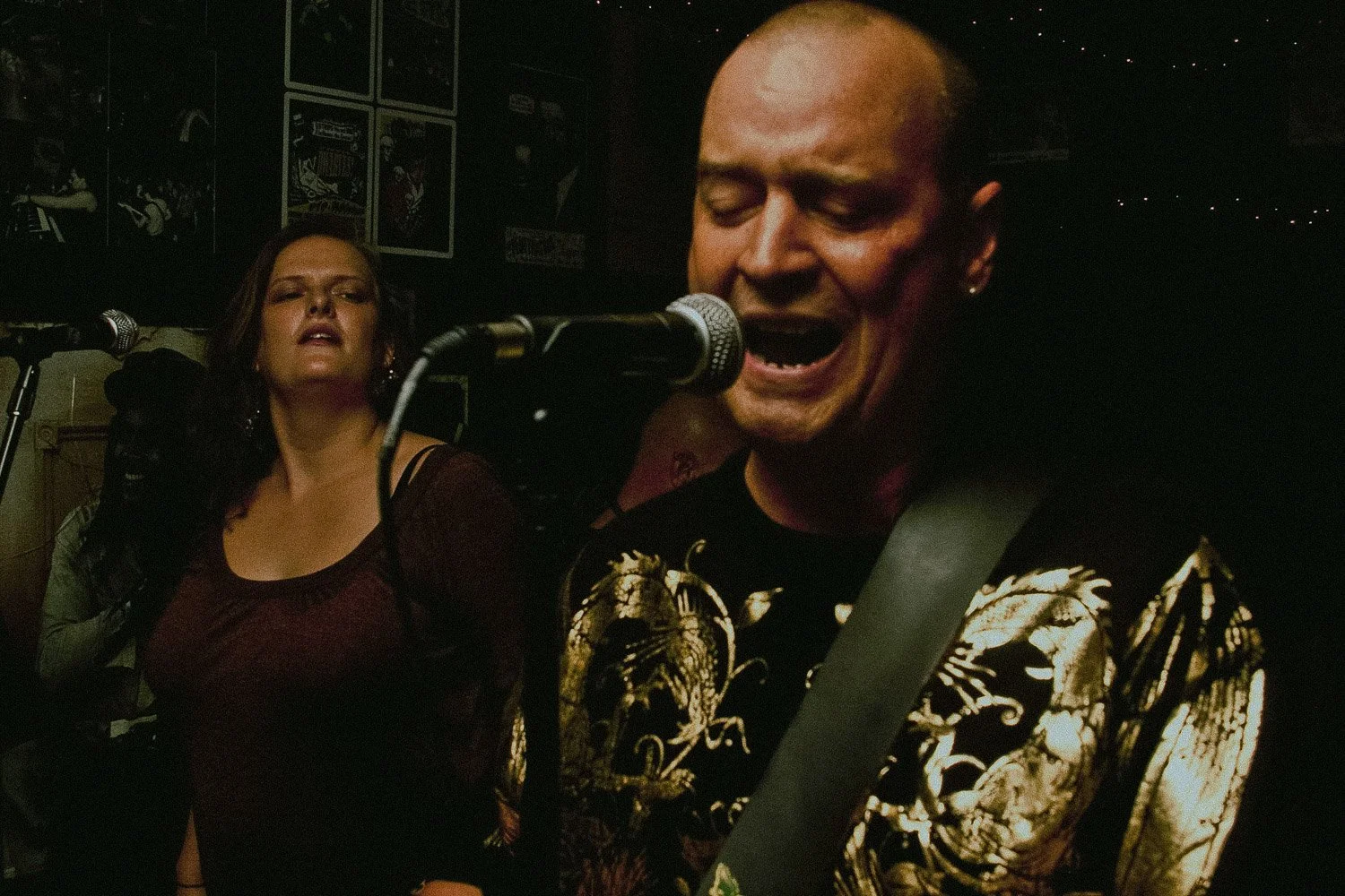 A man singing passionately into a microphone with a woman in the background in a dimly lit venue decorated with framed posters.