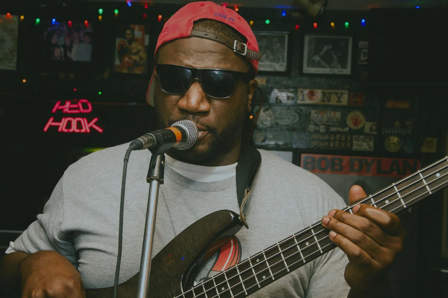 A man wearing sunglasses and a red cap playing an electric bass guitar and singing into a microphone in a dimly lit bar or music venue, with colorful string lights and vintage music posters and signs in the background.