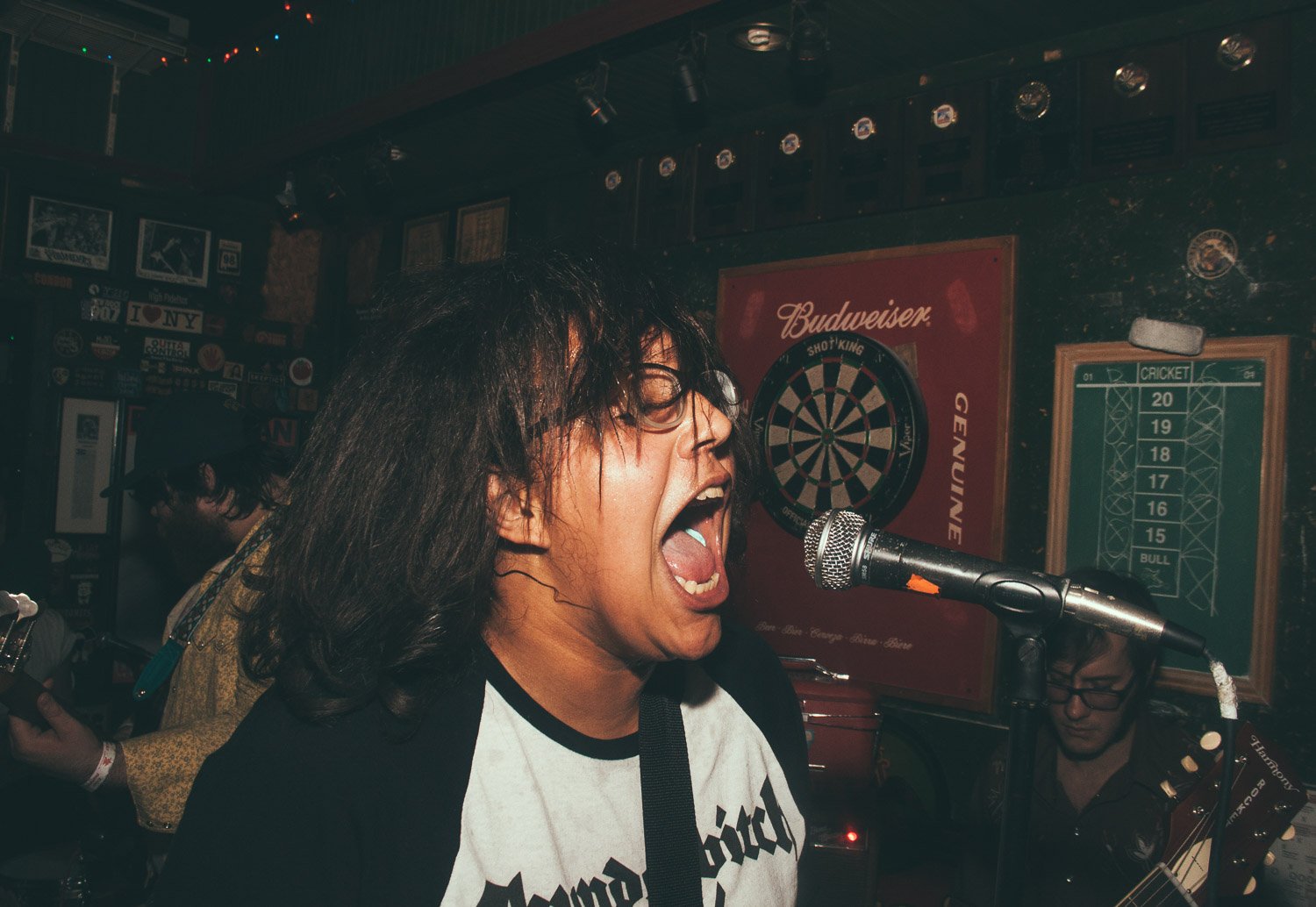 A woman singing passionately into a microphone at a bar or pub with dartboards and wall decorations in the background.