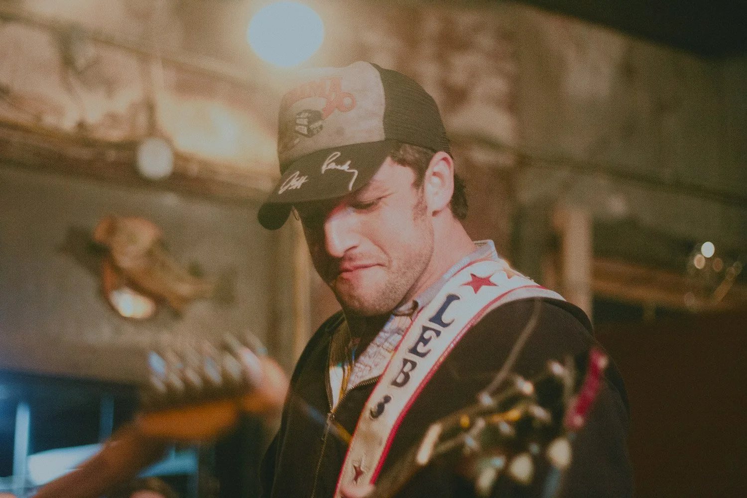 A man with a guitar, wearing a cap and a jacket with a guitar strap, smiles with closed eyes in a rustic indoor setting with warm lighting.