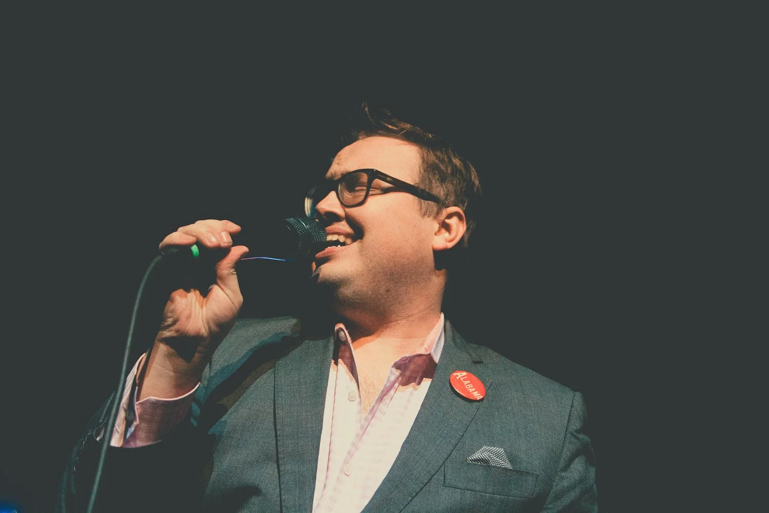 A man wearing glasses and a suit jacket, smiling while speaking into a microphone, with a red Alabama pin on his lapel.