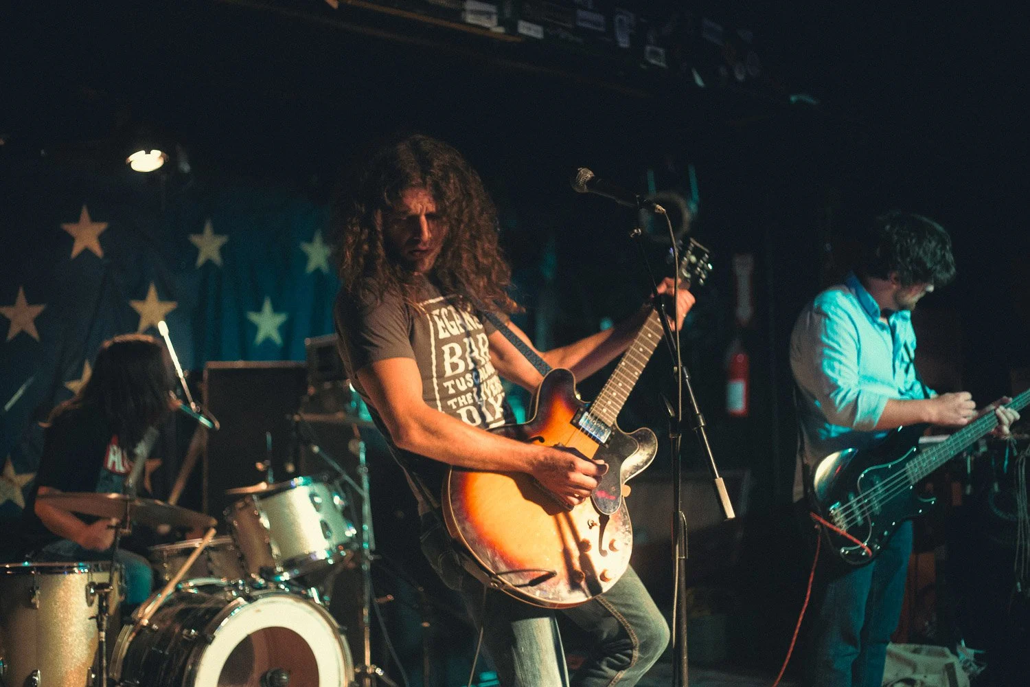 Band performing live on stage with a drummer, a guitarist, and a bassist, in front of a star-patterned backdrop.