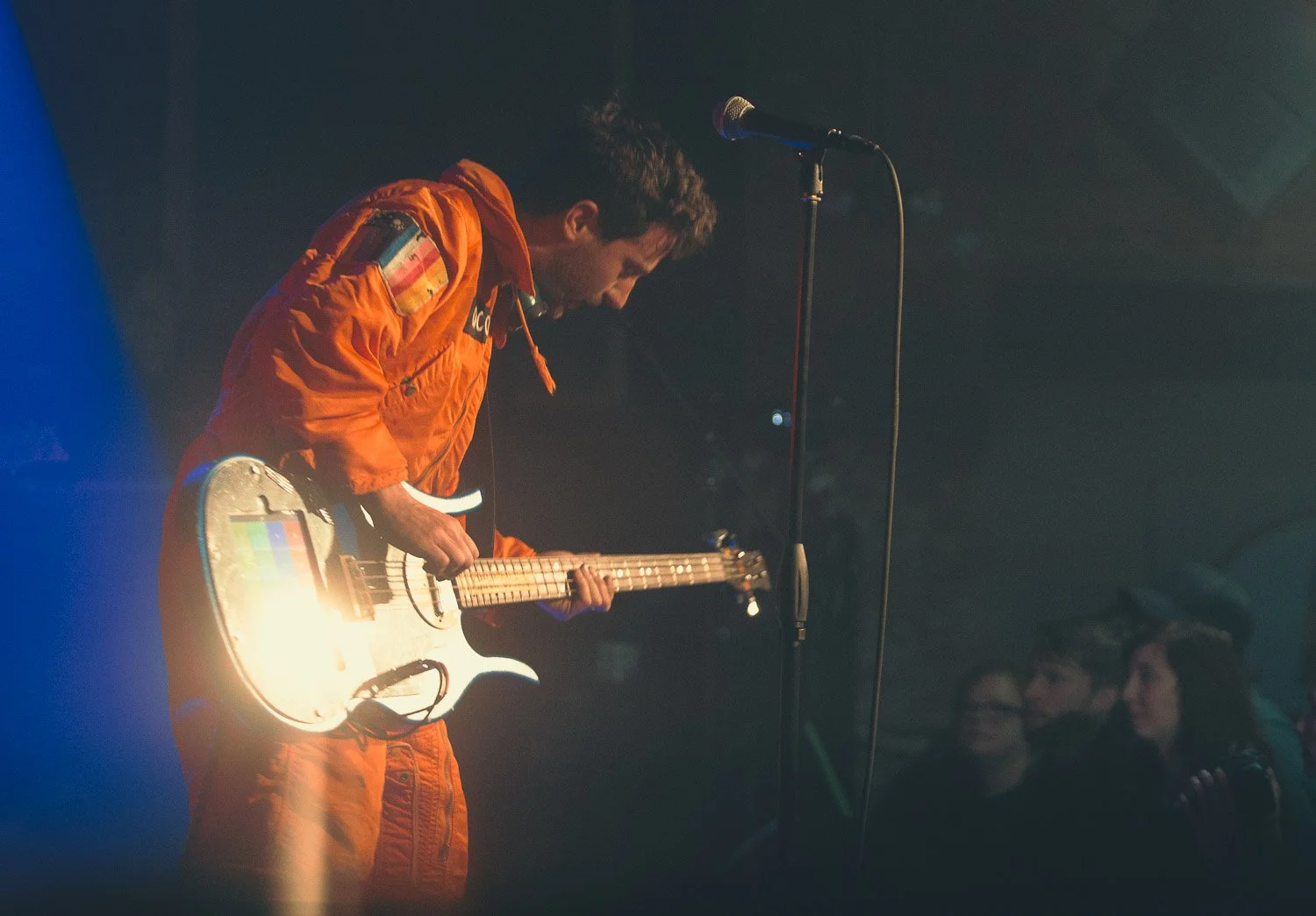 A musician dressed in an orange space suit playing an electric guitar on stage, with a microphone nearby and a small crowd watching in the background.