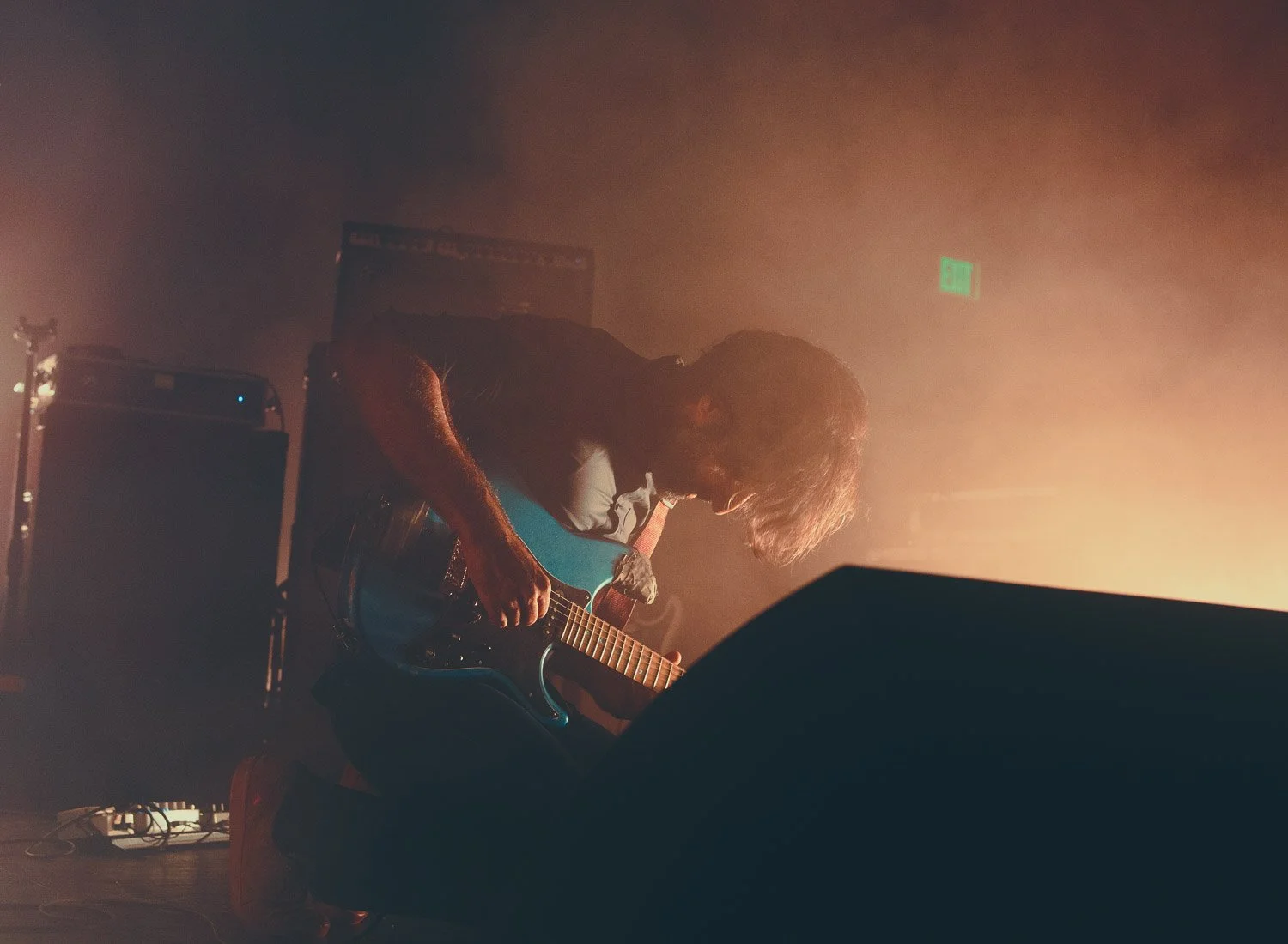 Musician with long hair playing blue electric guitar on stage with warm lighting.