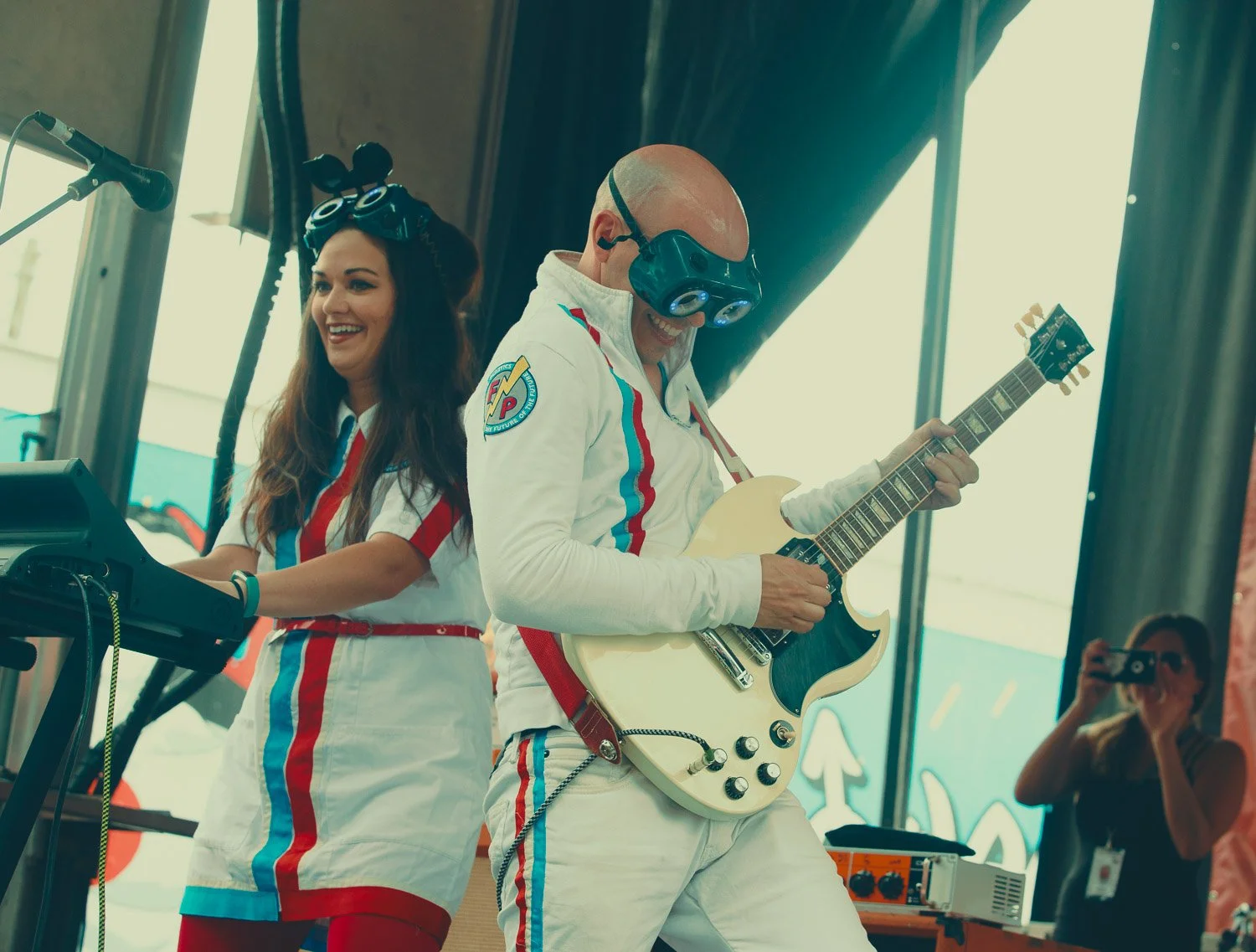 Two musicians in retro racing suits performing on stage, with one playing guitar and the other singing or playing keyboard, in a lively indoor setting.