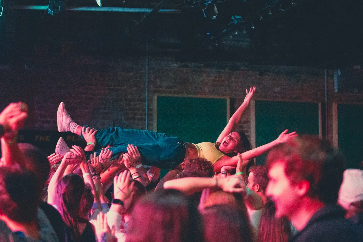 A woman being crowd-surfed at a concert, lying on people's hands with her arms raised, in a lively indoor venue with a brick wall background.