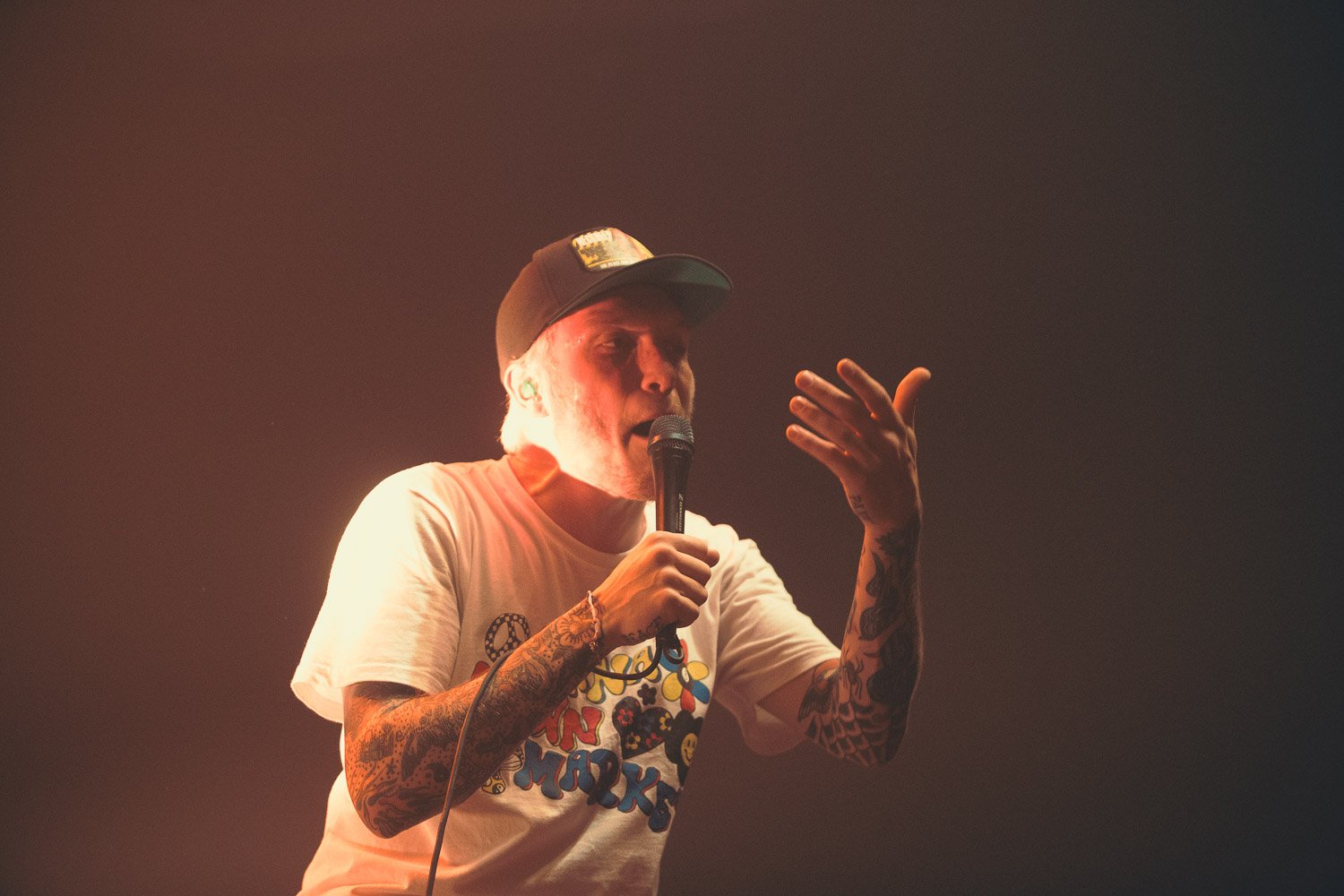A man with tattoos on his arms, wearing a white t-shirt with colorful graphics and a black baseball cap, holding a microphone and speaking on stage with reddish lighting.