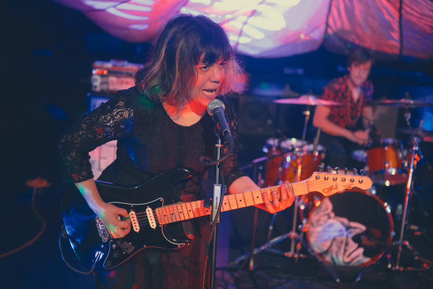 Female musician singing into microphone and playing electric guitar on stage, with drummer in background at a live music venue.
