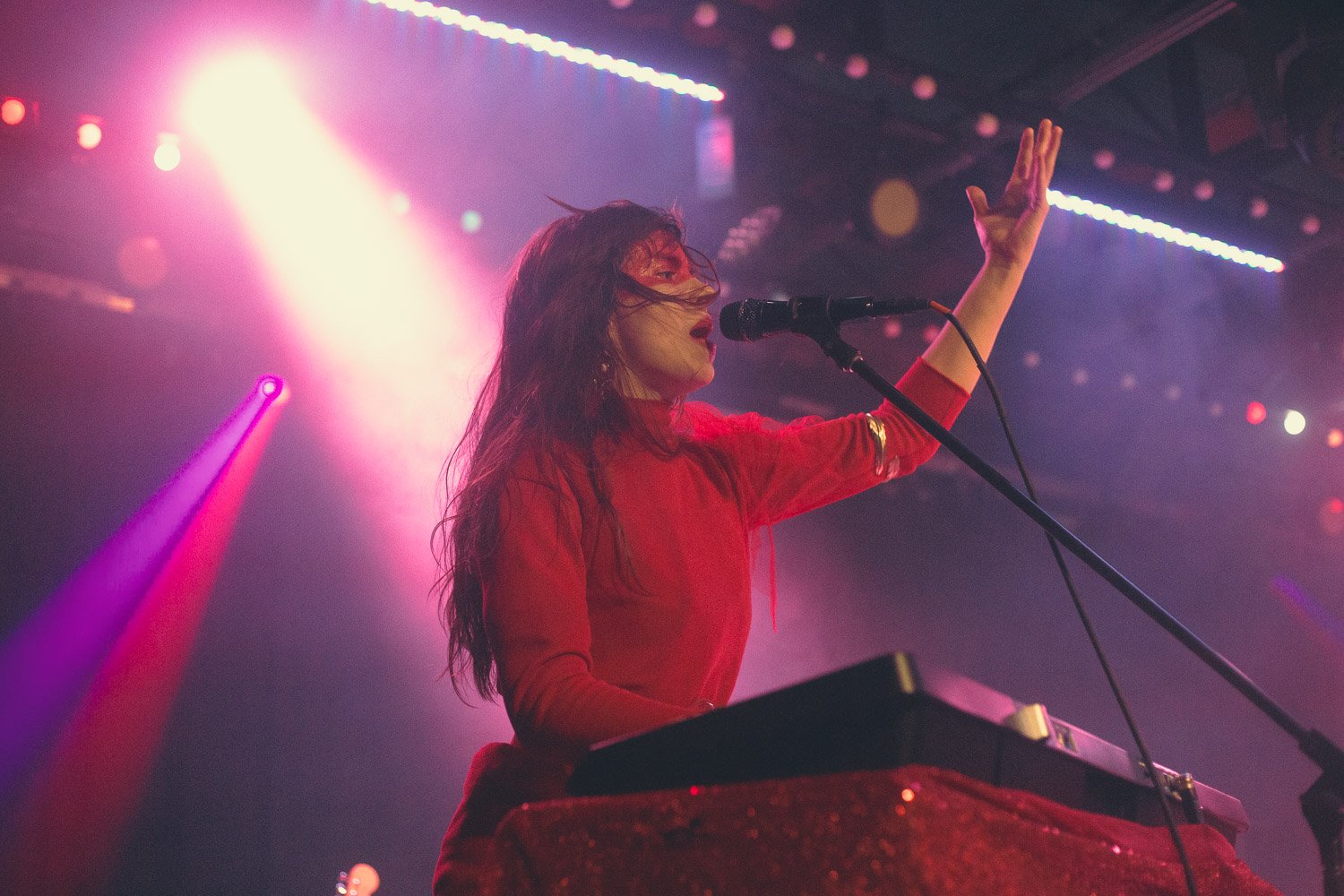 A female musician singing into a microphone while playing a keyboard on stage, with colorful stage lighting and fog creating a vibrant atmosphere.