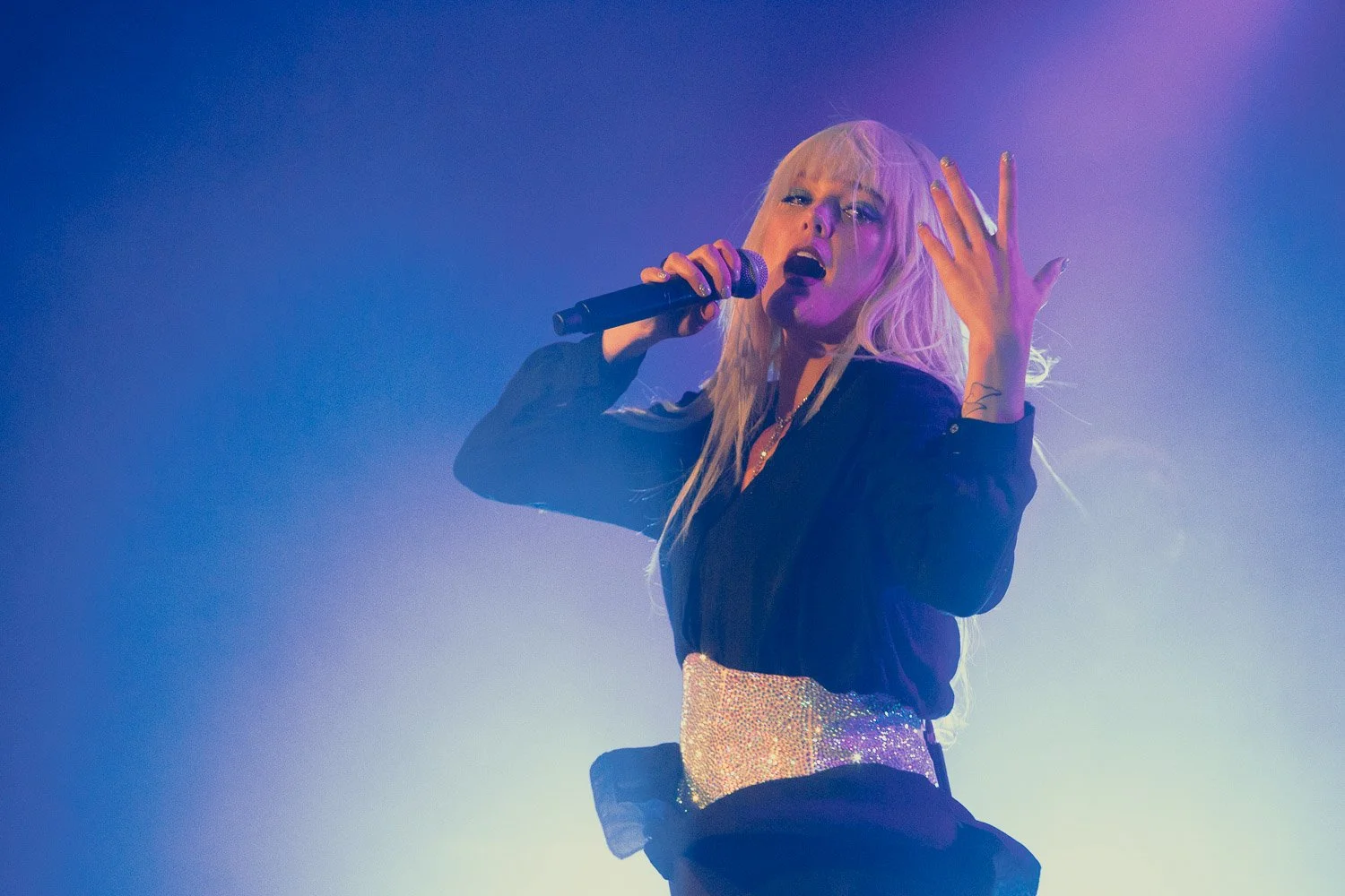 A blonde female singer performing on stage, holding a microphone in her right hand, with her left hand raised, against colorful stage lighting.
