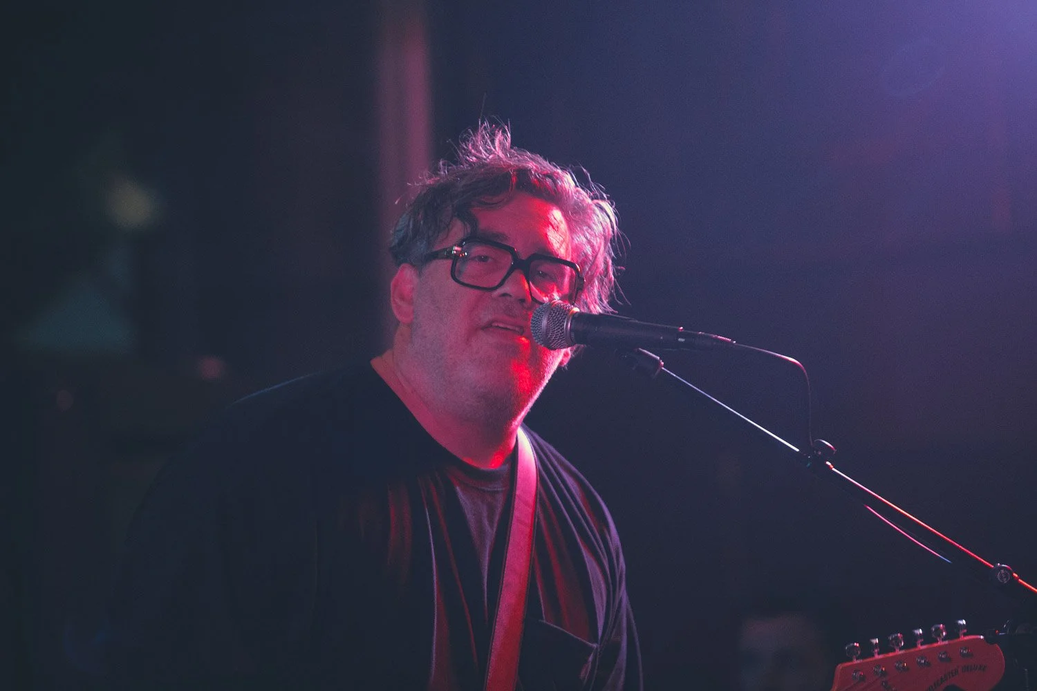 A man with glasses singing into a microphone on stage, playing a guitar under pink and purple stage lighting.