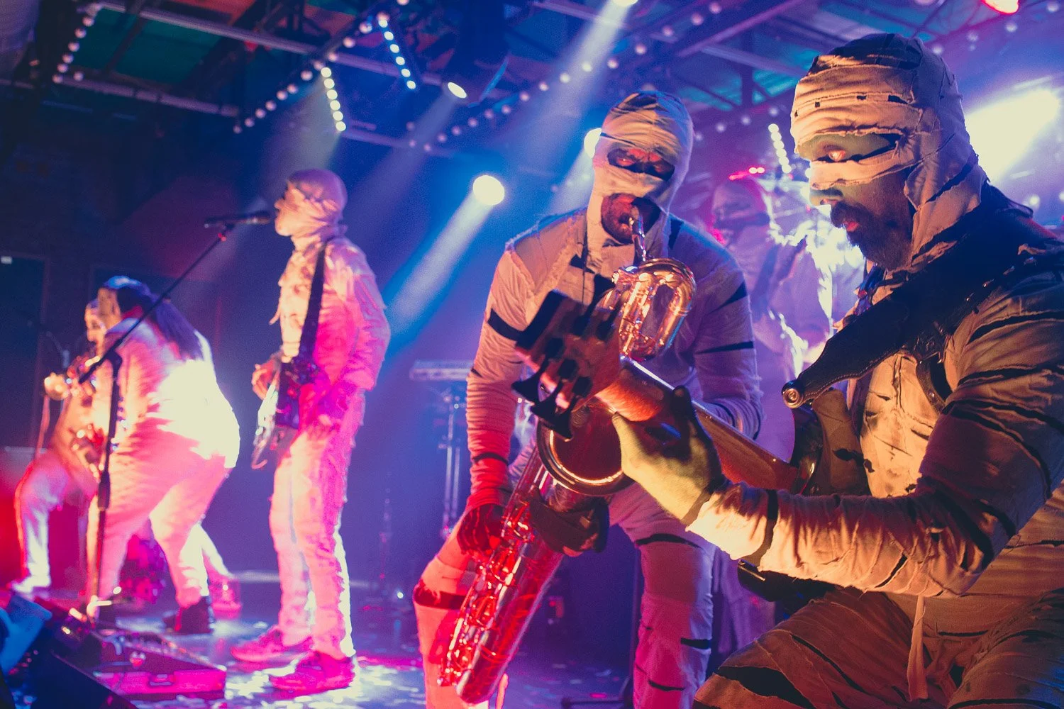 Band performing on stage with five members in coordinated beige outfits with head wraps, playing various instruments under colorful stage lighting.