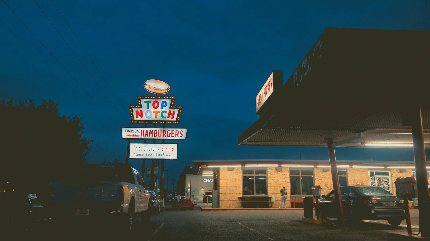 Nighttime view of a fast food restaurant with a colorful illuminated sign reading 'Top Notch', offering charcoal hamburgers, fried chicken, and shrimp, with parking lot and cars in front.