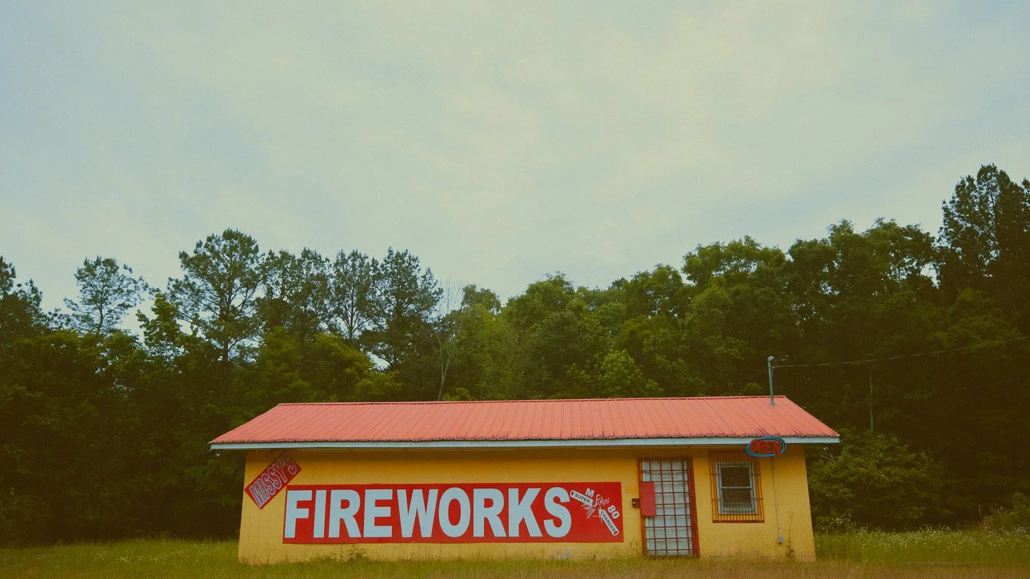 A small yellow building with a red roof and sign that reads 'FIREWORKS' in large white letters. Additional signs say 'Missy' and 'Open'. Trees in the background and a grassy area in front.