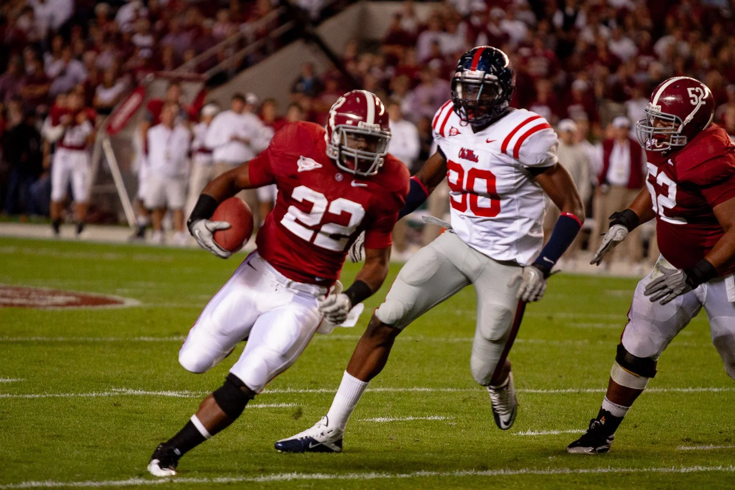 College football game with players in red and white uniforms on the field, one player in red carrying the ball while others defend.