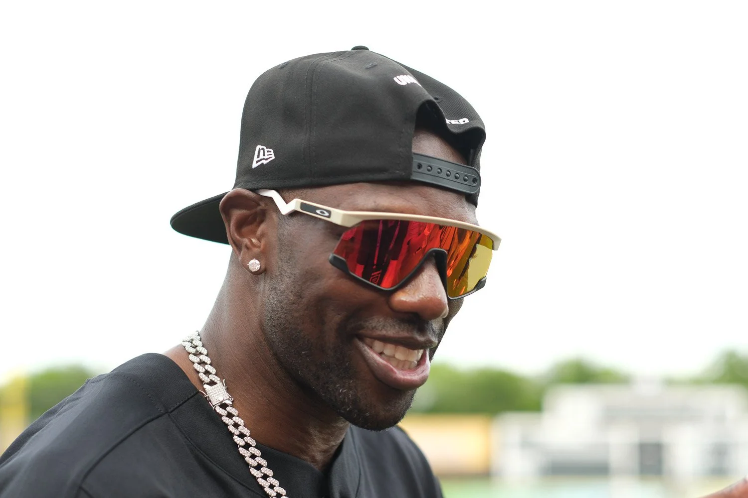 Close-up of a man wearing a black cap backwards, orange-tinted sports sunglasses, diamond earrings, a chain necklace, and a black shirt, smiling outdoors.