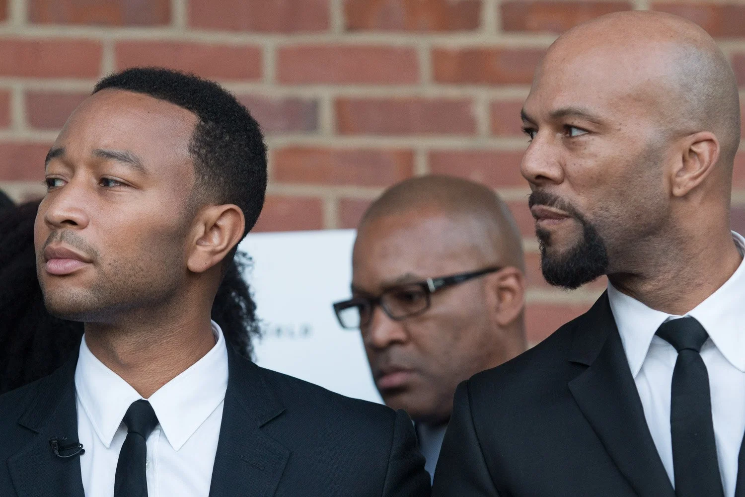 Three men dressed in formal black suits, white shirts, and black ties, standing outside with a brick wall in the background. The man on the left has short dark hair, the man on the right is bald with facial hair, and the man in the middle wears glass
