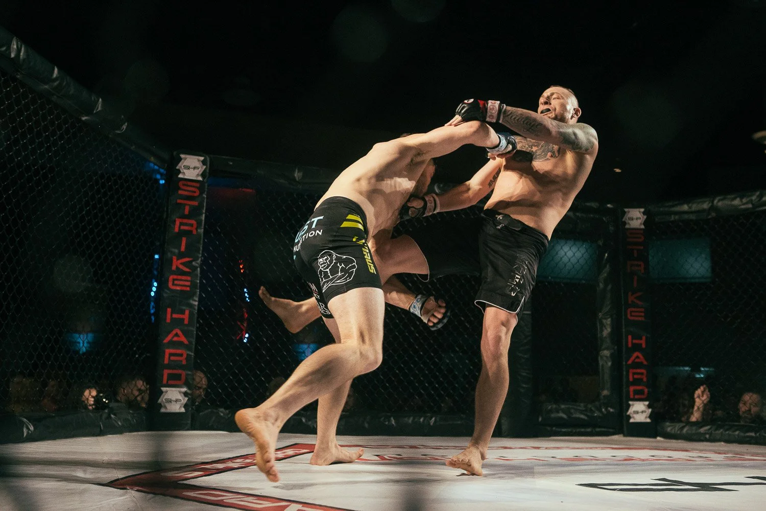Two mixed martial arts fighters inside an octagon cage, one delivering a punch to the other's face. Fighter on the left is wearing black shorts with yellow and white accents, while fighter on the right is wearing black shorts and has tattoos on his a