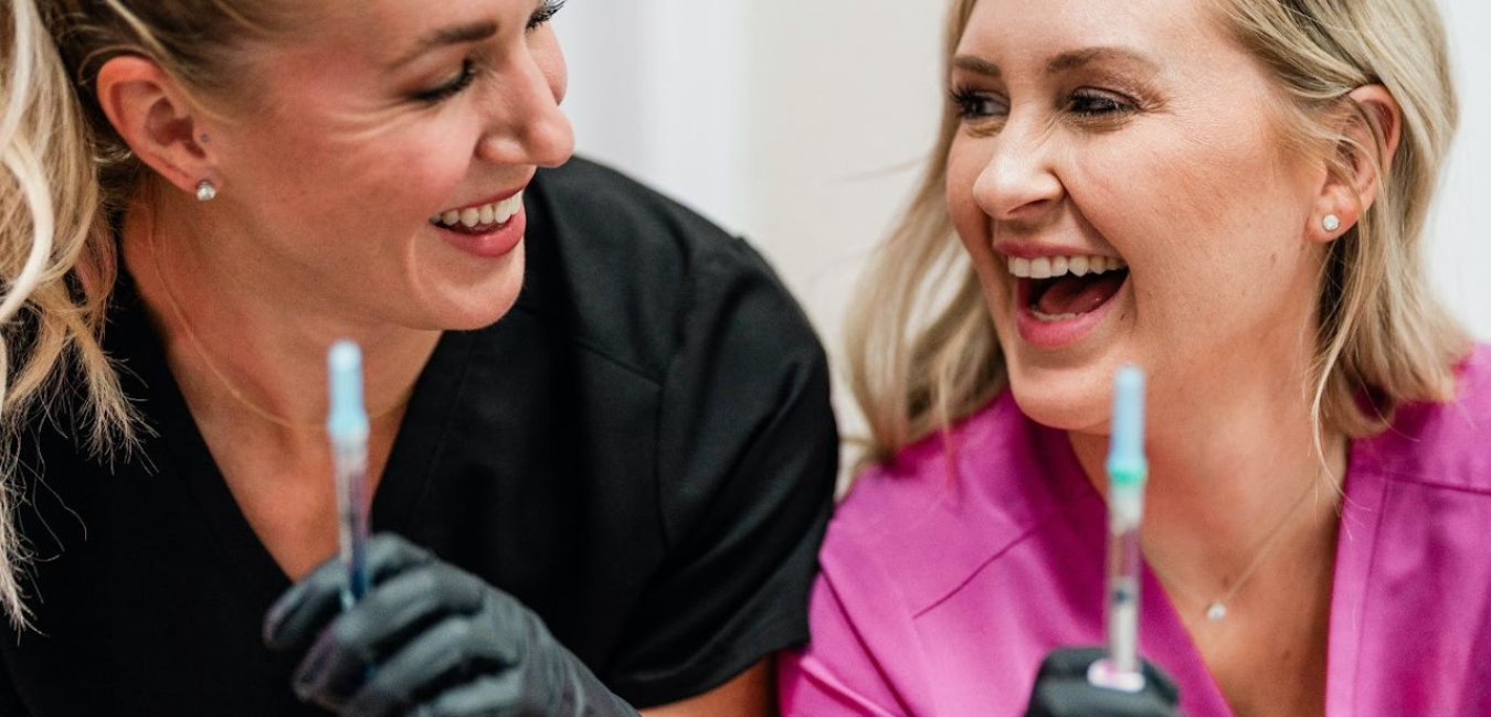 Two female medical professionals in scrubs smiling and laughing while holding syringes.