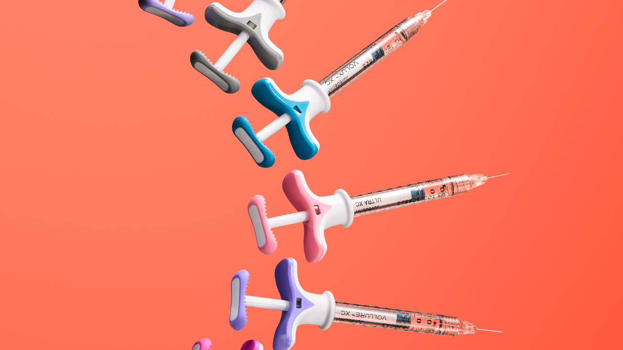 Multiple colorful syringes with nozzles pointed downward against a coral background.