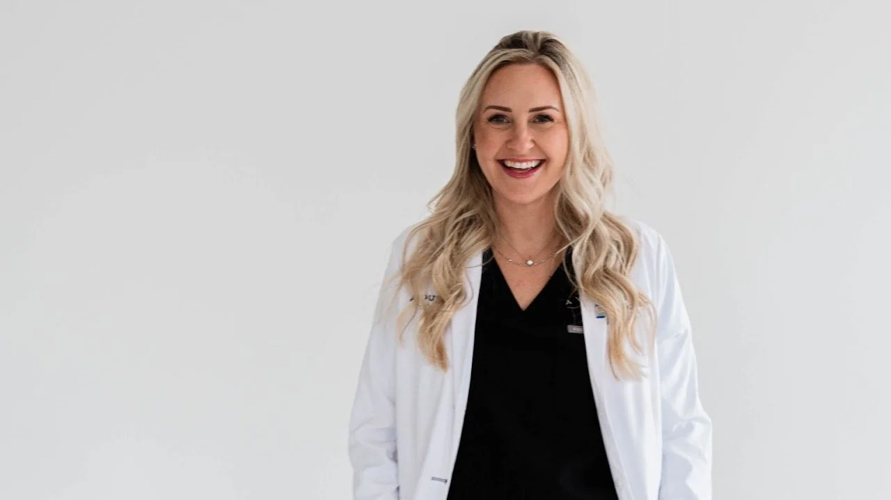 A smiling woman with long blonde hair, wearing a black top and white lab coat, standing against a plain white background.