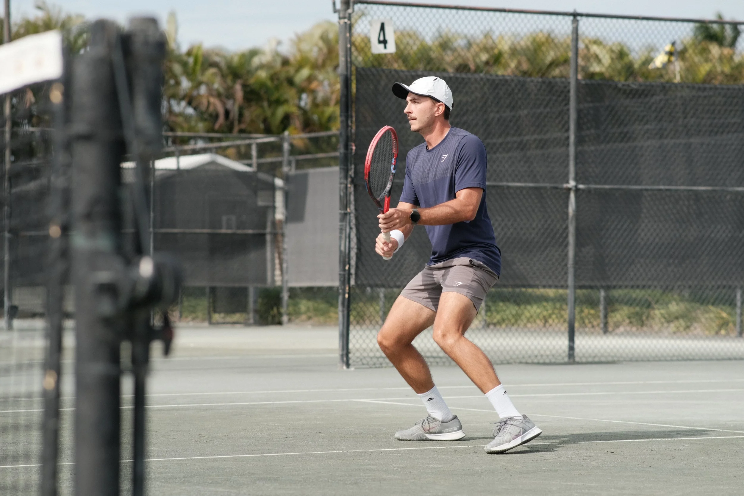 A man playing tennis on an outdoor court, preparing to hit a shot with a tennis racket, wearing sportswear and a white cap.