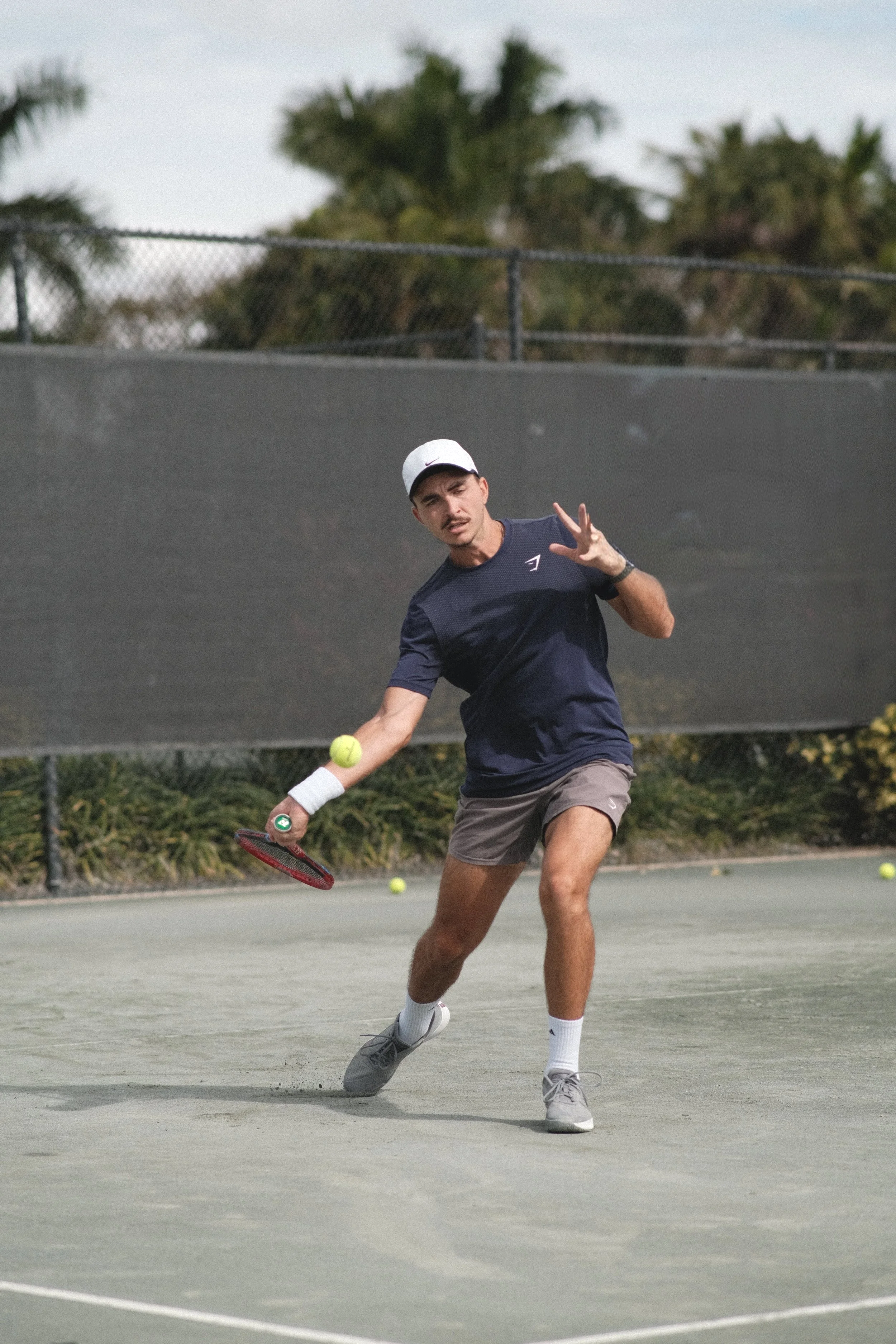 Young man playing tennis on an outdoor court, wearing a white cap and navy shirt, preparing to hit a tennis ball with a racquet.