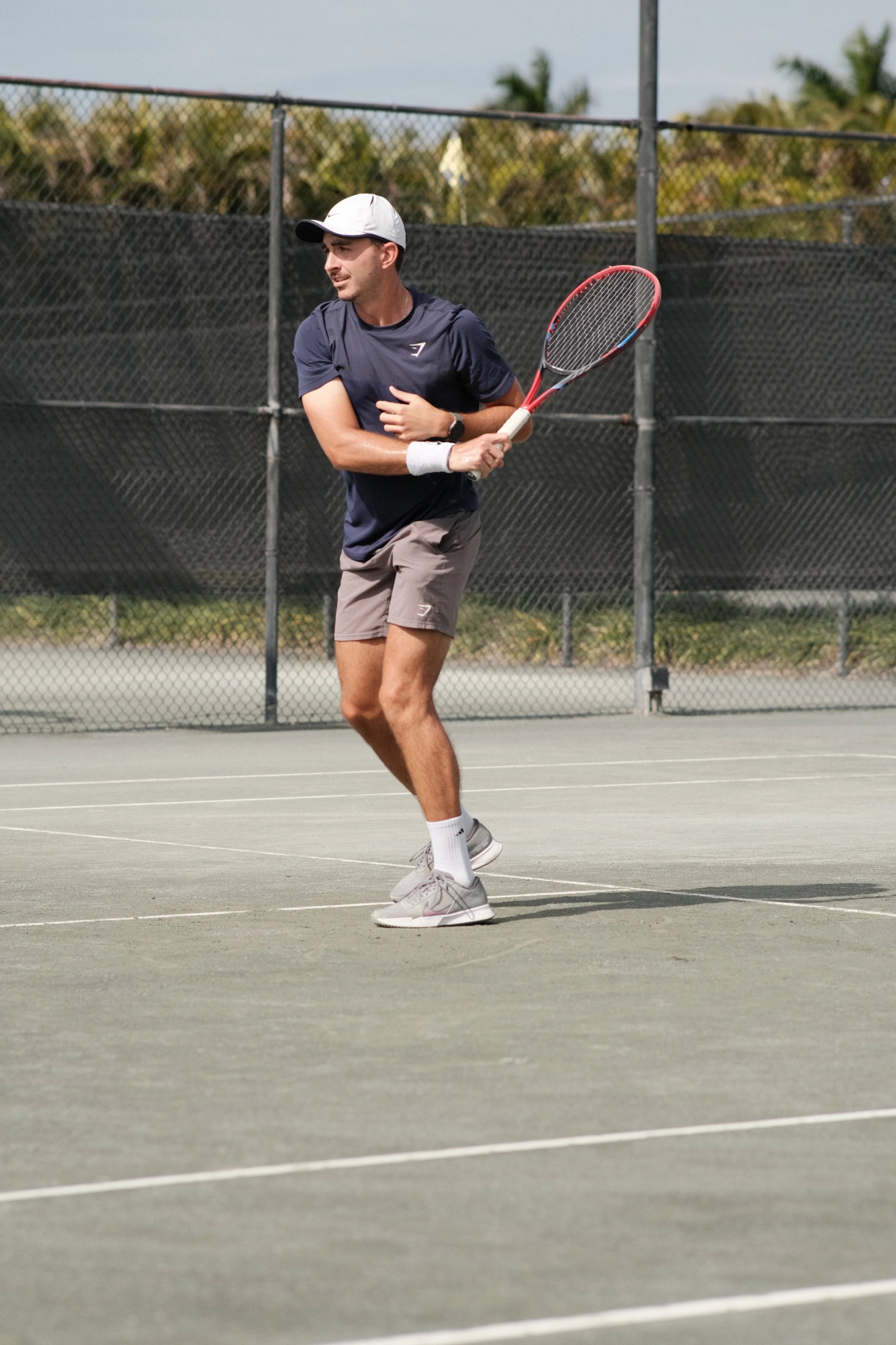 A man playing tennis on an outdoor court, holding a tennis racket, wearing a white cap, navy shirt, grey shorts, and grey sneakers.