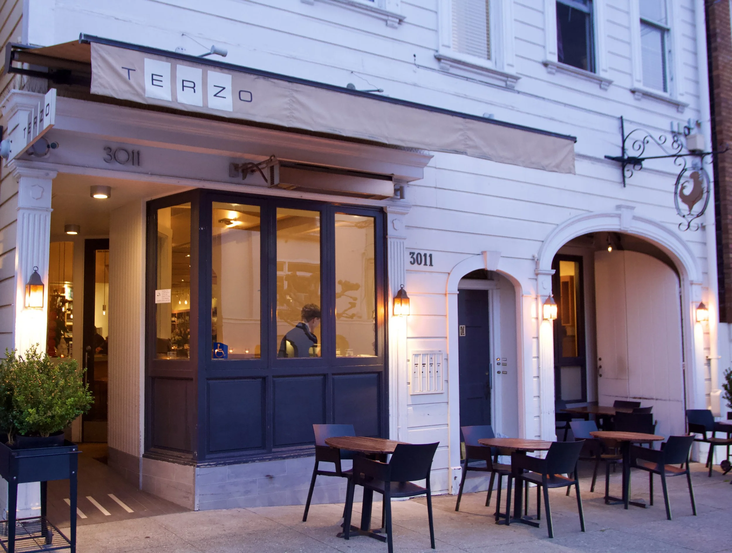 Exterior of a restaurant named Terzo at 3011 with outdoor seating, black chairs, small wooden tables, and lighting fixtures on white walls.