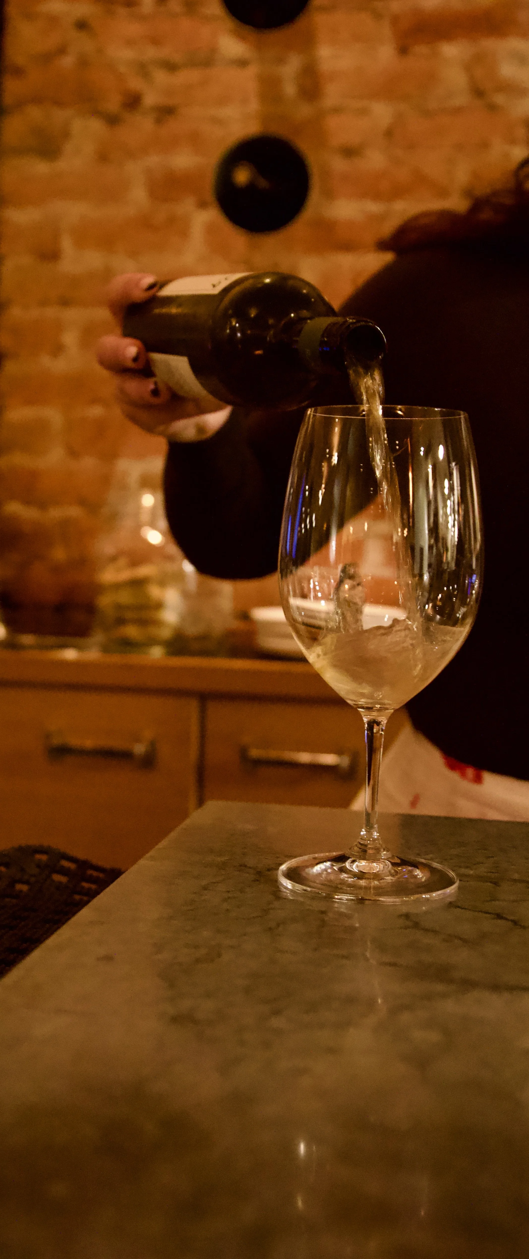 A person pouring a drink into a wine glass at a bar with a brick wall background.