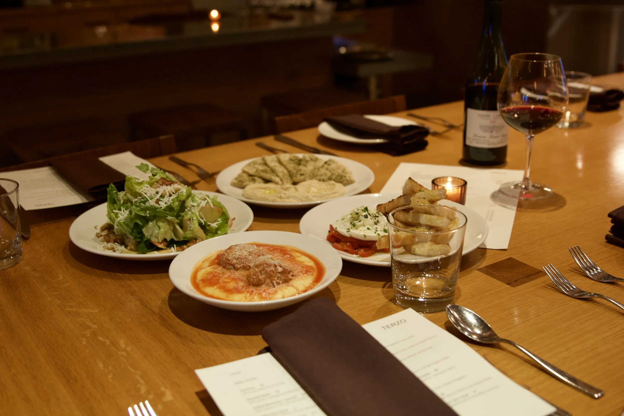 A wooden dining table set with various Italian dishes including a salad, pizza, meatballs in tomato sauce, a plate with bread and cheese, a poached egg with prosciutto, and a dish with French fries. There is a glass of red wine, a bottle of wine, a l