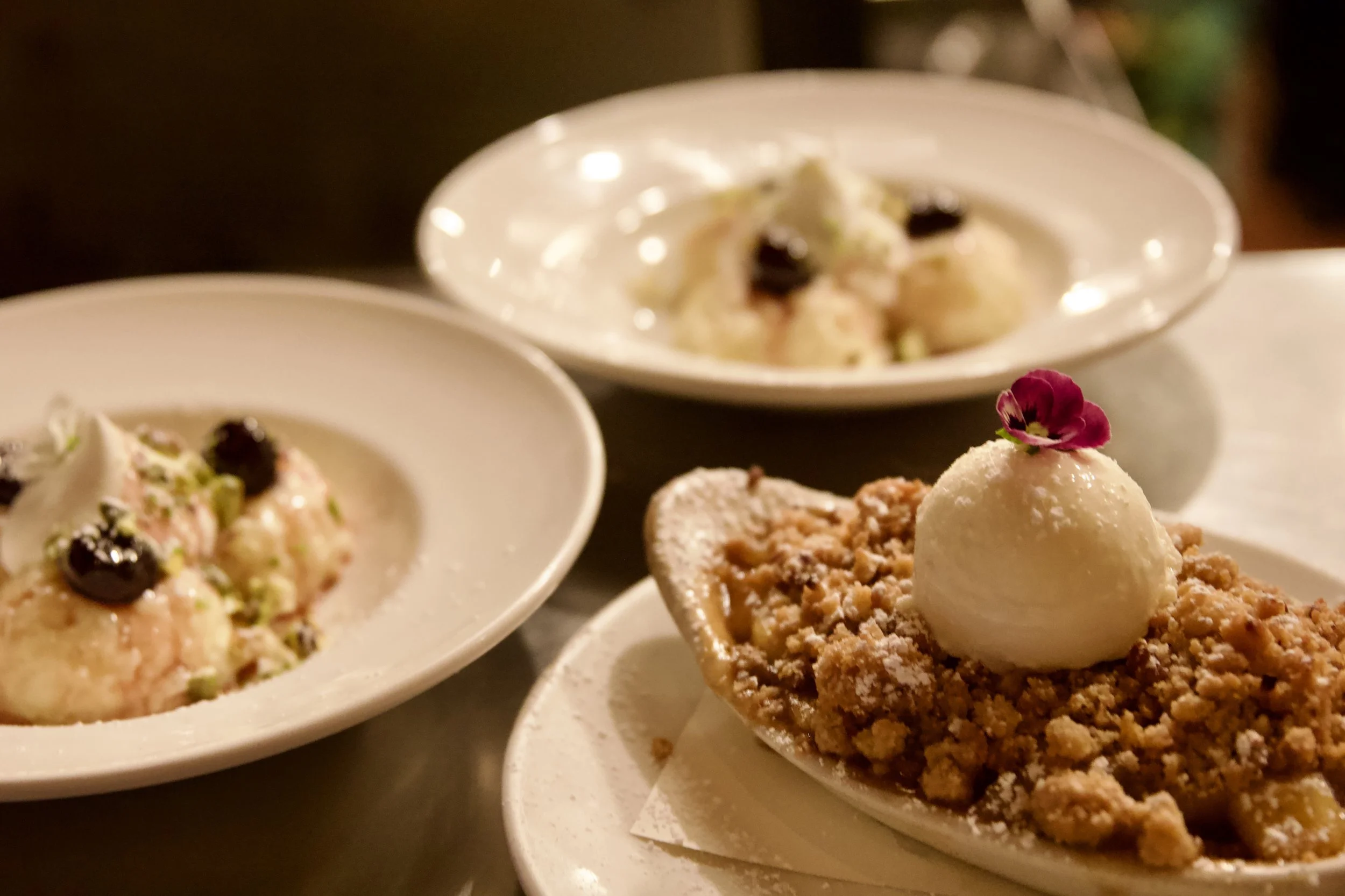 Three plates of desserts, with one prominently featuring a crumbly topping, a scoop of vanilla ice cream, and a small flower garnish; two other plates with creamy desserts topped with blueberries and other fruits.
