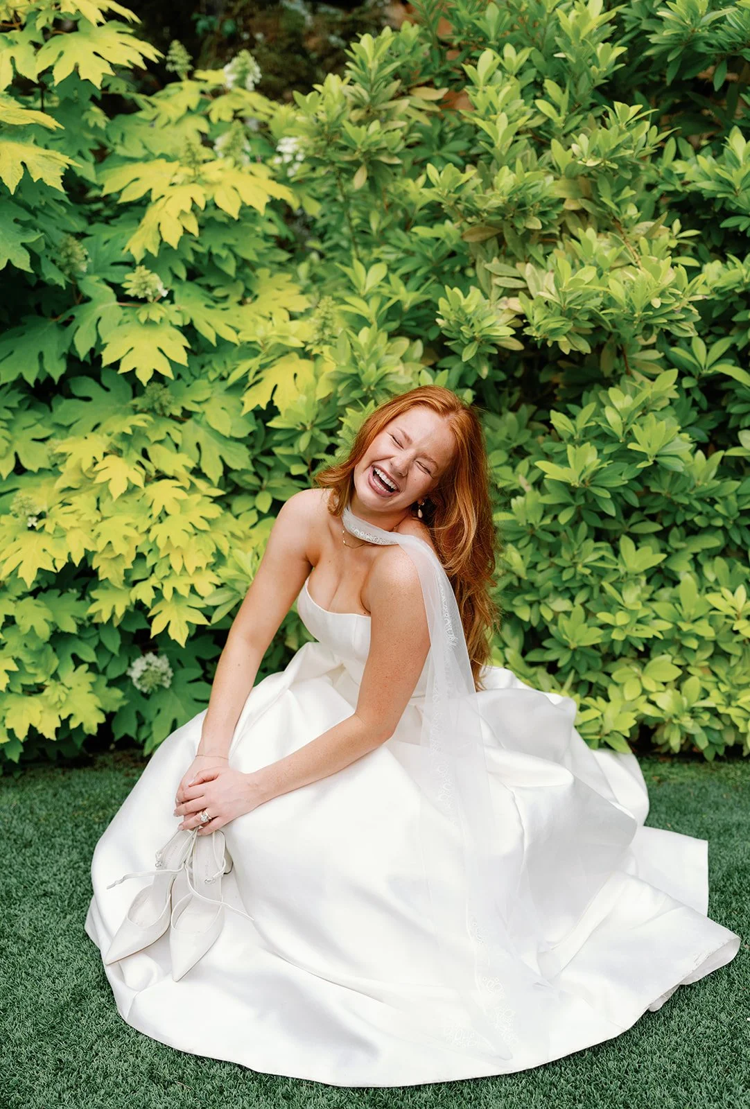 A smiling bride in a wedding dress sits on grass in front of green bushes, holding her shoes.