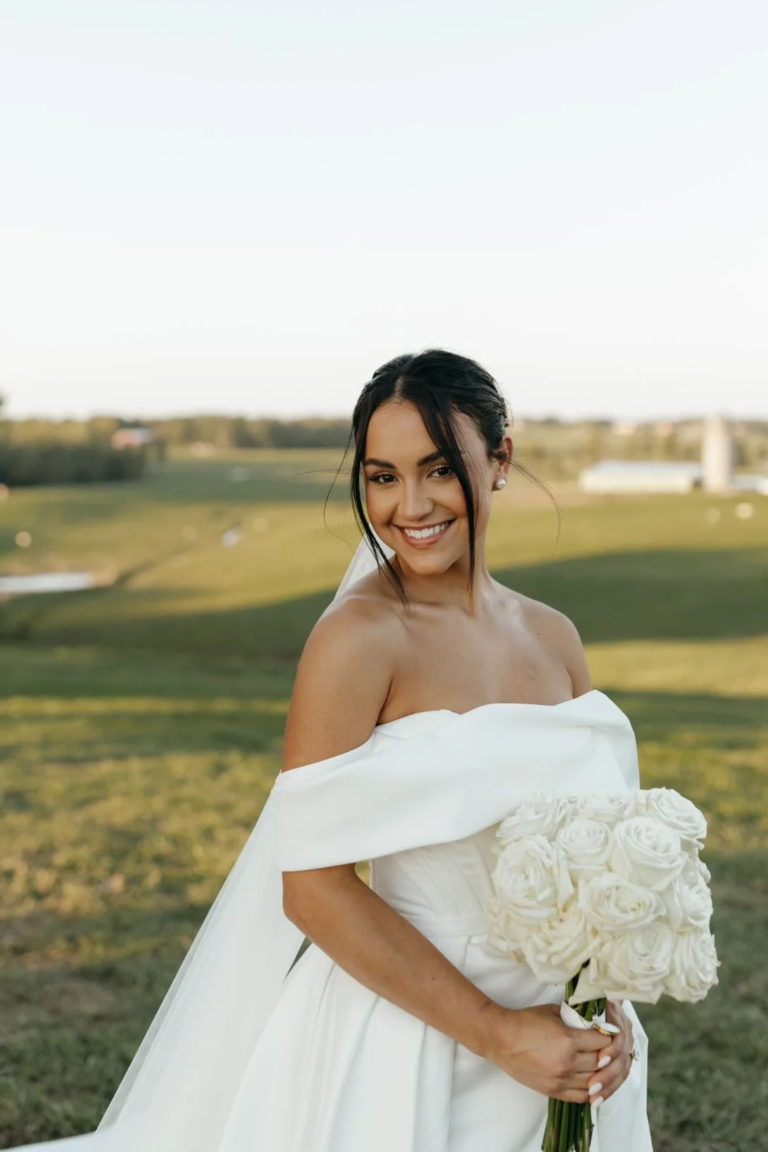 A smiling young woman in a white off-shoulder wedding dress holding a bouquet of white roses, standing outdoors in a grassy area under a clear sky.